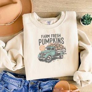 以下が含まれることがあります： 「FARM FRESH PUMPKINS」の文字と、カボチャでいっぱいのヴィンテージのティールトラックのグラフィックがプリントされたクリーム色のスウェットシャツ。スウェットシャツは、ブルージーンズ、茶色の帽子、サンダルと組み合わせています。