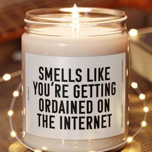 May include: A white candle with a black label that reads "Smells like you're getting ordained on the internet". The candle is lit and sitting on a wooden coaster.