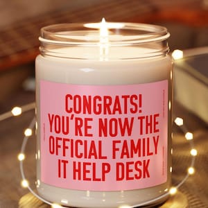 May include: A white candle in a glass jar with a pink label that reads "Congrats! You're now the official family IT help desk". The candle is lit and sitting on a wooden coaster.