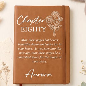 May include: A tan notebook with an elastic closure and a ribbon bookmark. The cover features white text reading Chapter EIGHTY, a floral line drawing, a printed quote, and the name Aurora in a script font centered at the bottom of the cover.