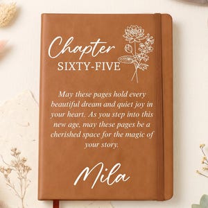 May include: A tan leather-bound journal featuring a white floral line drawing, the text Chapter SIXTY-FIVE, a printed quote about dreams and joy, and the name Mila in script. The notebook includes a vertical elastic band and a red ribbon bookmark.