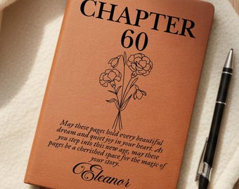 May include: A brown leather journal with the text "CHAPTER 60" and a floral illustration. The journal also includes personalized text and a ribbon bookmark. A black pen rests beside the journal.