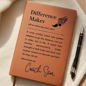 May include: A brown leather-bound notebook with the words "Difference Maker" and a definition of a cross country coach. The notebook has a black pen and a red bookmark. The notebook is on a white blanket.