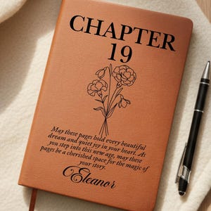 May include: A brown leather journal with the text "CHAPTER 19" and a floral illustration. The journal includes a personalized message and the name "Eleanor". A black pen rests beside the journal.