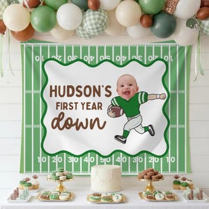 May include: A festive banner with the text "HUDSON'S FIRST YEAR down" and a cartoon illustration of a baby playing football. The banner is set against a green and white football field backdrop, with a balloon arch above.