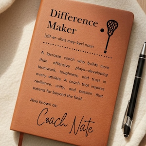 May include: A brown leather-bound notebook with the words "Difference Maker" and a lacrosse stick graphic. The text defines a lacrosse coach and is signed "Coach Nate". A black pen rests beside the notebook.