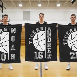 May include: Three black blankets with a basketball theme, featuring white text and graphics. Each blanket displays a basketball design, the word "TIGERS", a name, a number, and a letter. The blankets are held by three people in a gymnasium.