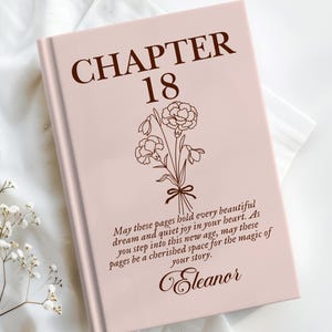 May include: A blush-colored book with the text "CHAPTER 18" in brown. A floral illustration and a quote are below the title. The book is on a white surface with floral accents.