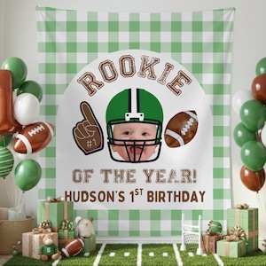 May include: A birthday party scene with a green and white checkered backdrop. The backdrop features a football-themed graphic with the text "Rookie of the Year!" and "Hudson's 1st Birthday." Balloons in green, brown, and white decorate the scene, along with gift boxes and a football-themed rug.