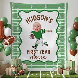 May include: A festive first birthday party scene with a football theme. A backdrop features a cartoon illustration of a baby playing football, with the text "HUDSON'S FIRST YEAR down". Balloons in green, white, and brown complete the decor.