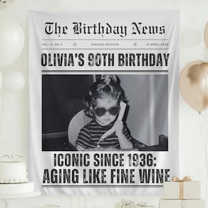 May include: A white fabric banner resembling a newspaper front page, celebrating "OLIVIA'S 90TH BIRTHDAY." The banner includes a black and white photo of a young girl wearing heart-shaped sunglasses. The text reads "ICONIC SINCE 1936: AGING LIKE FINE WINE."