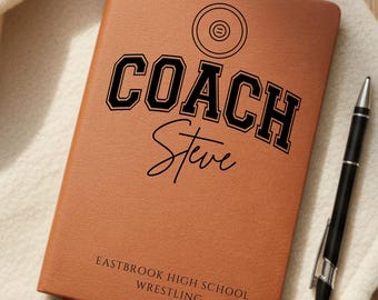 May include: A brown leather notebook with the words "COACH Steve" in black lettering. The notebook also features the text "EASTBROOK HIGH SCHOOL WRESTLING" and a pen resting on a white cloth.