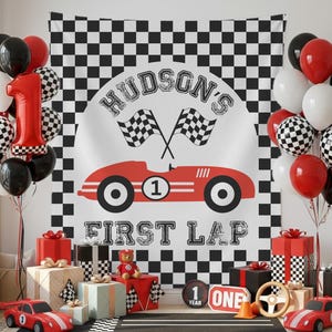 May include: A racing-themed first birthday party setup with a checkered backdrop and balloons. The banner reads "Hudson's First Lap" with a red race car illustration. Red, black, white, and checkered balloons, gifts, and toy race cars complete the decor.