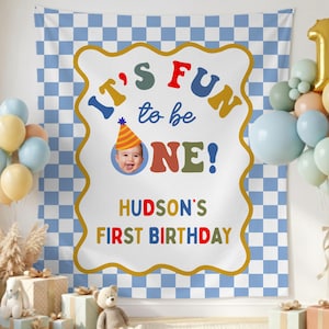 May include: A blue and white checkered backdrop with a large sign that reads "It's Fun to be ONE!" with a photo of a baby. The sign also says "Hudson's First Birthday". Balloons and gifts are in the foreground.