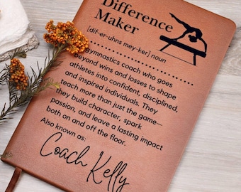 May include: A brown leather journal with the words "Difference Maker" and a definition. The journal includes a gymnast silhouette and the words "Coach Kelly" at the bottom. A sprig of orange flowers rests on the journal.