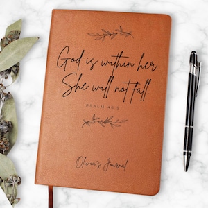 May include: A brown journal with the text "God is within her. She will not fall. Psalm 46:5" and "Olivia's Journal" in black script. A black and silver pen rests on the right side of the journal. Decorative leaves are in the background.