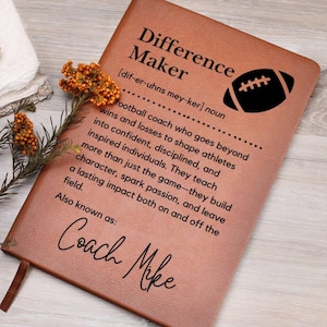 May include: A brown leather-bound notebook with the words "Difference Maker" and a football graphic. The definition of a football coach is printed below. The notebook is personalized with the name "Coach Mike".