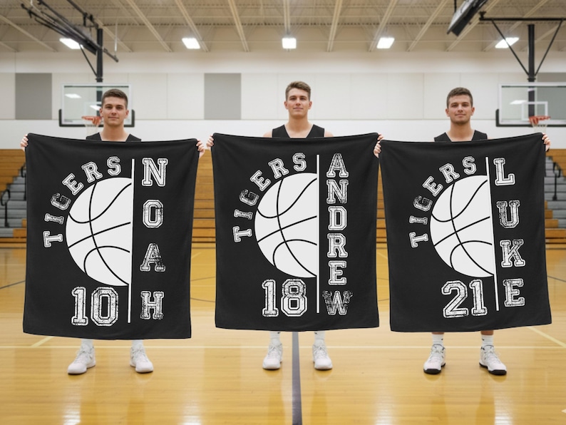 May include: Three black blankets with a basketball theme, featuring white text and graphics. Each blanket displays a basketball design, the word "TIGERS", a name, a number, and a letter. The blankets are held by three people in a gymnasium.