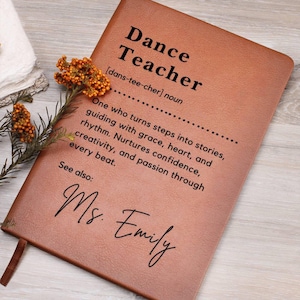May include: A brown leather-bound notebook with the words "Dance Teacher" and a definition printed on the cover. The notebook includes the name "Ms. Emily" and a decorative floral element.