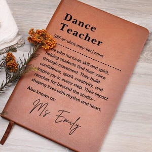 May include: A brown leather-bound notebook with the words "Dance Teacher" and a definition printed in black. The notebook also includes the name "Ms. Emily" in script. A sprig of orange flowers rests on the corner.