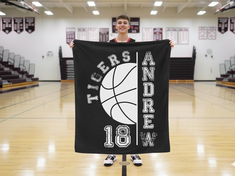 May include: Black blanket with a white basketball graphic, the word "TIGERS" above and "18W" below. The name "ANDREW" is printed vertically on the right. The blanket is held in front of a person in a gymnasium.