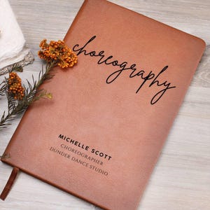 May include: A brown leather-bound notebook with the word "choreography" in cursive script. The notebook also features the name "Michelle Scott," the title "Choreographer," and "Dunder Dance Studio." A sprig of orange flowers rests on the cover.