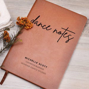 May include: A brown leather-bound notebook with the words "dance notes" in black script. The notebook also includes the name "Michelle Scott", the title "Choreographer", and "Dunder Dance Studio". A brown ribbon bookmark is visible.