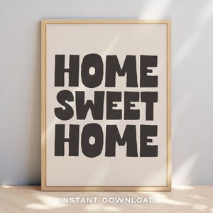 Puede incluir: Un diseño de arte de pared imprimible con el texto "Home Sweet Home" en letras negras y negritas sobre un fondo blanco. El diseño está enmarcado en un marco de madera claro.