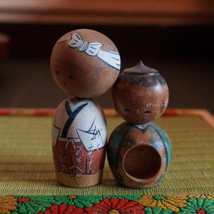 以下が含まれることがあります： 白いヘッドバンドをした伝統的な日本の木製のドールと、緑と茶色の模様の服を着たもう1つのドール。ドールは顔の表情が描かれており、織られたマットの上に立っています。