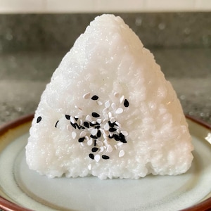 Könnte beinhalten: Ein dreieckiger Onigiri, oder Reisball, liegt auf einem kleinen Teller. Der weiße Reis ist mit schwarzen und weißen Sesamsamen bestreut. Der Teller hat eine hellgrüne Mitte und einen braunen Rand.
