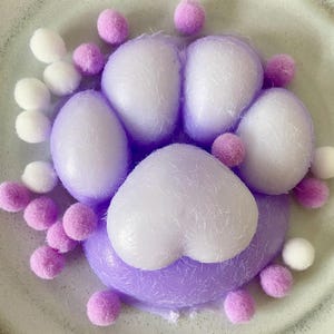 Peut inclure: Une empreinte de patte décorative dans les tons lavande et blanc, entourée de petits pompons duveteux. L'empreinte de patte est centrée sur une assiette en céramique de couleur claire, créant une présentation fantaisiste et artistique.