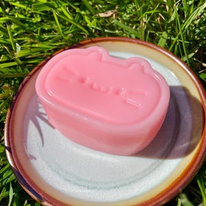 Könnte beinhalten: Ein rosa, katzenförmiges Seifenstück liegt auf einem kleinen, runden Keramikgeschirr mit braunem Rand. Die Seife hat eine glatte Oberfläche mit geprägten Gesichtszügen. Das Geschirr hat einen gesprenkelten, cremefarbenen Innenraum.