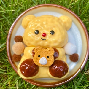 May include: A yellow bear-shaped omelet with a red nose and brown sauce on a small brown plate. The omelet is decorated with a small yellow bear-shaped cookie.