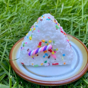 May include: A white, triangular cake slice with colourful sprinkles and a swirled candle on a small plate with a brown rim. The plate sits on green grass, creating a vibrant contrast.