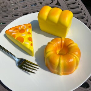 May include: A white plate holds a slice of pizza, a loaf of bread, and a bundt cake. The pizza slice is yellow with orange spots. The bread is yellow, and the bundt cake is orange. A silver fork is on the plate.