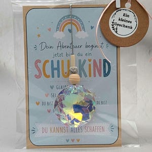 Könnte beinhalten: Ein Geschenkset mit einer Karte mit dem Text "Dein Abenteuer beginnt" und "SCHUKIND", einem Regenbogen und einem Kristallprismen-Ornament. Das Set enthält ein kleines Holzschild mit der Aufschrift "Ein kleines Geschenk".