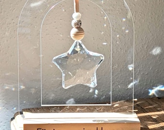 May include: A clear crystal star ornament suspended within a clear acrylic arch, resting on a wooden base. The base is engraved with the German phrase "Für immer im Herzen." Decorative wooden beads hang above the star.