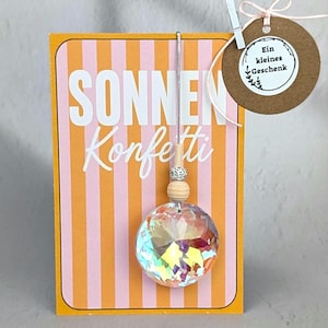 Könnte beinhalten: Eine rechteckige Karte mit vertikalen orangefarbenen und rosafarbenen Streifen und dem Text SONNEN Konfetti. Ein runder, irisierender Kristall hängt an einer Schnur mit zwei Holzperlen. Ein braunes, rundes Etikett mit dem Text Ein kleines Geschenk ist befestigt.