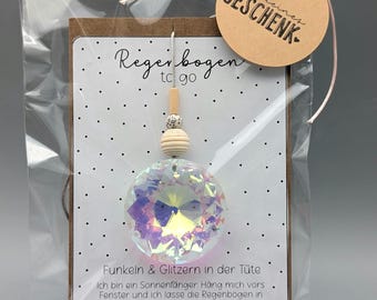 May include: A clear, faceted crystal suncatcher with iridescent colors, packaged in a clear bag with a brown tag and a small wooden clip. The tag reads "a small gift" in German. The card inside says "Rainbow to go".