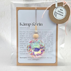 Könnte beinhalten: Ein klarer, facettierter Kristallanhänger mit einer Holzperle und einem kleinen Anhänger mit der Aufschrift "Ein kleines Geschenk". Der Anhänger hängt vor einer Karte mit deutschem Text.