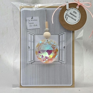 Könnte beinhalten: Eine kleine Geschenkkarte mit einer dekorativen Kristallkugel, die vor einer Fensterillustration hängt. Die Karte trägt den Text "Ein bisschen Sonne für dich" und ein braunes Etikett mit dem Text "Ein kleines Geschenk". Die Karte ist in einer transparenten Plastiktüte verpackt.
