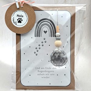 Könnte beinhalten: Eine Grußkarte mit Regenbogen-Design, Pfotenabdrücken und dem Text "Und am Ende des Regenbogens sehen wir uns wieder." Ein Holzanhänger mit dem Namen "Nala" und einem Pfotenabdruck ist befestigt.