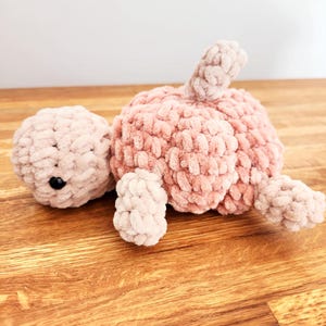 Può includere: Un giocattolo di tartaruga rosa all'uncinetto con una texture soffice. La tartaruga è sdraiata sulla schiena con le gambe e la testa distese.