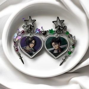Könnte beinhalten: Zwei herzförmige Acryl-Charms mit Anime-Charakteren. Jedes Charm zeigt ein anderes Bild. Die Charms sind durch eine Kette aus Perlen, Sternen und Schmetterlingen verbunden. Kleine silberne Dolche hängen von unten.