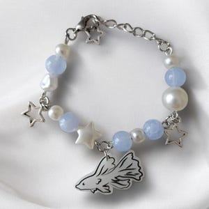 K&ouml;nnte beinhalten: Ein silbernes Armband mit Sternanh&auml;ngern, hellblauen Perlen und wei&szlig;en Perlen. Ein Fisch-Charm h&auml;ngt am Armband. Das Armband wird auf einer wei&szlig;en, strukturierten Oberfl&auml;che pr&auml;sentiert.