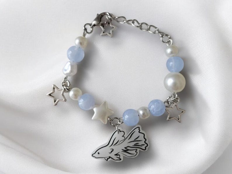 K&ouml;nnte beinhalten: Ein silbernes Armband mit Sternanh&auml;ngern, hellblauen Perlen und wei&szlig;en Perlen. Ein Fisch-Charm h&auml;ngt am Armband. Das Armband wird auf einer wei&szlig;en, strukturierten Oberfl&auml;che pr&auml;sentiert.
