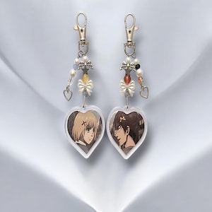 Op de afbeelding: Twee hartvormige sleutelhangers met anime-personageportretten. Elke sleutelhanger heeft een zilveren sluiting, kralenaccenten, een strik en een hartvormige bedel. De portretten tonen een blond personage en een bruinharig personage.