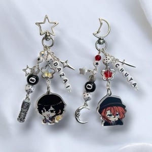 Peut inclure: Deux porte-clés argentés avec des breloques. Un porte-clés a un charme d'étoile et l'autre a un charme de croissant de lune. Les deux porte-clés ont des breloques avec les noms "DAZAI" et "CHUYA" dessus. Les porte-clés ont également des breloques de dés, d'étoiles et d'un téléphone.