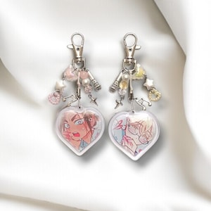 May include: A pair of heart-shaped keyrings with anime-style illustrations. Each keyring has a silver clasp, a microphone charm, and decorative beads. The heart pendants feature colourful character art.