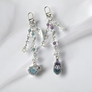 May include: Two silver charm keychains with colourful beads, stars, and flowers. One keychain features a cartoon character with blue hair and a red shirt, the other features a cartoon character with purple hair and a green hat.